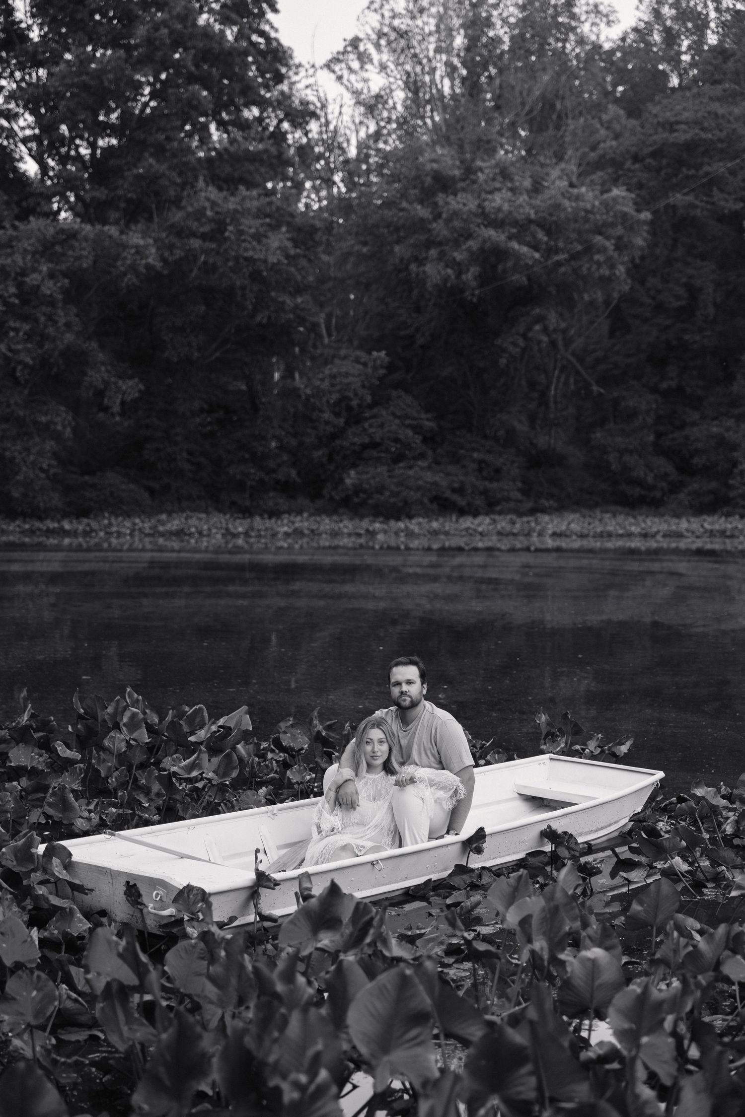 Black and white photo of a couple sitting together peacefully in a white boat on a lake with lily pads.