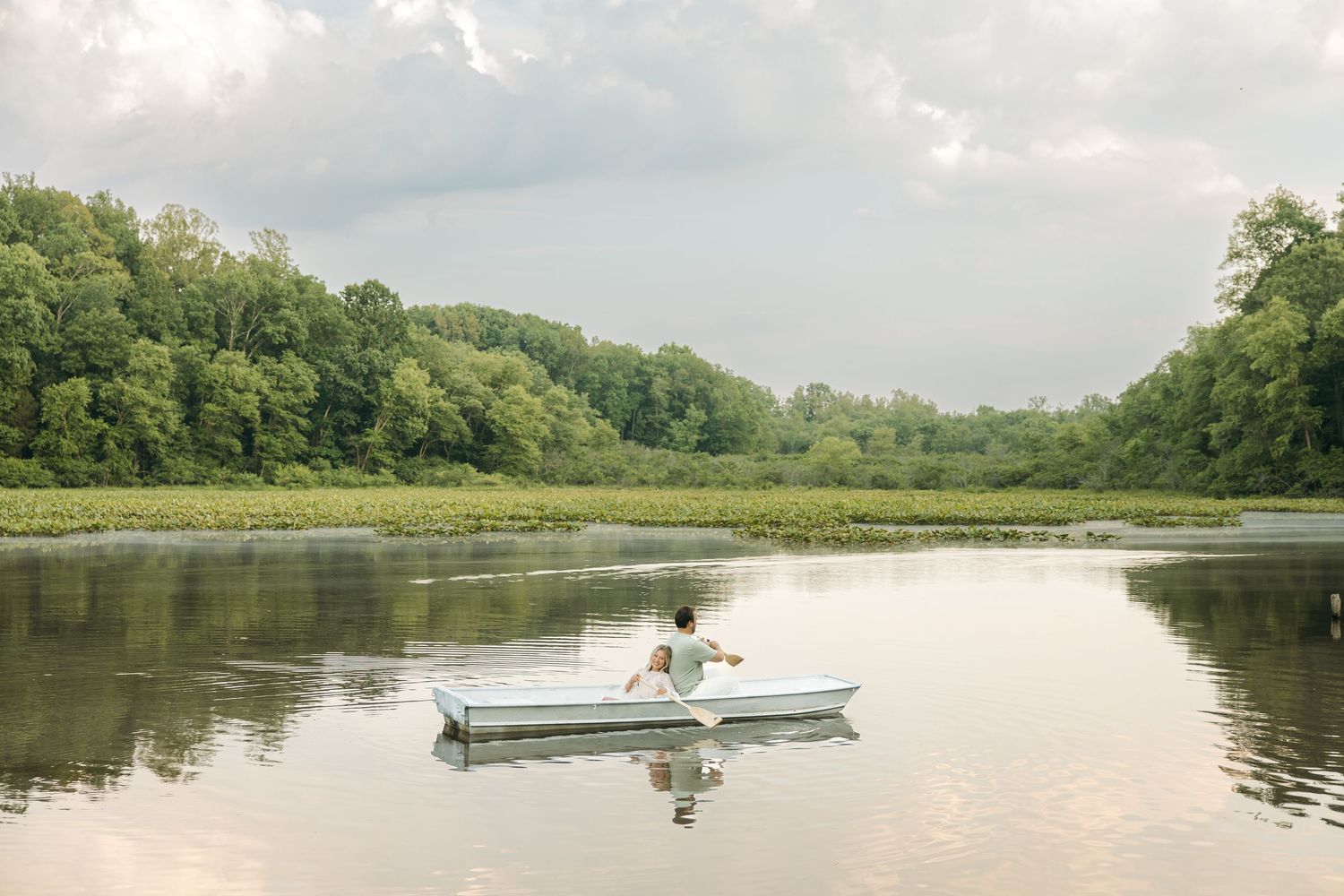 Person rowing a small white boat across a serene lake with green forested shoreline and cloudy sky reflected in water.