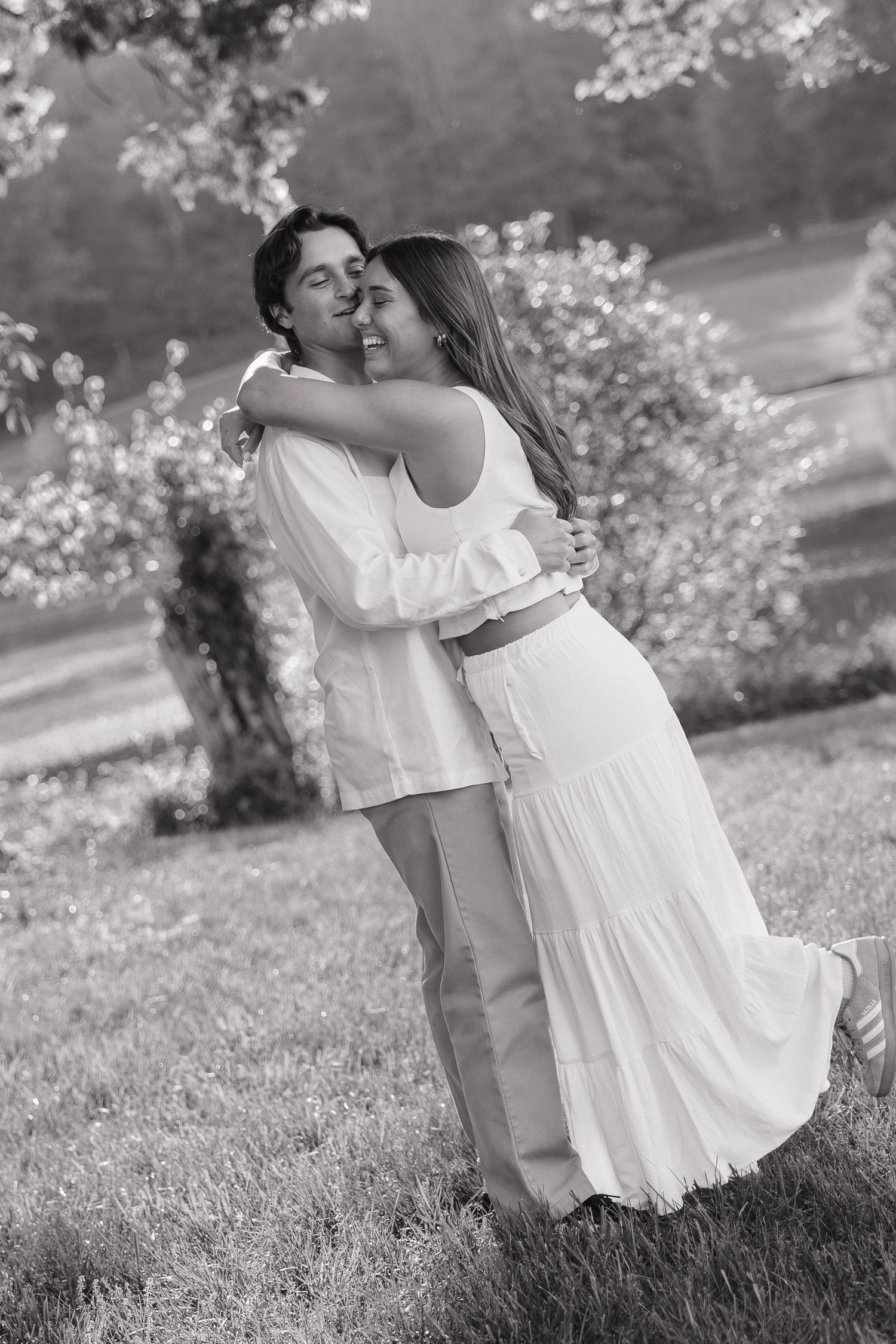 Black and white photo of a joyful couple embracing and laughing together on a green lawn near blooming trees.