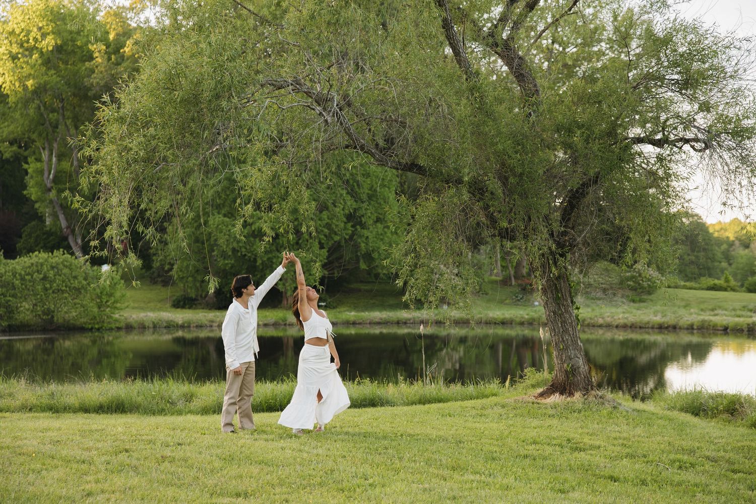 Couple dancing outdoors near a large willow tree beside a calm pond in a green park setting.