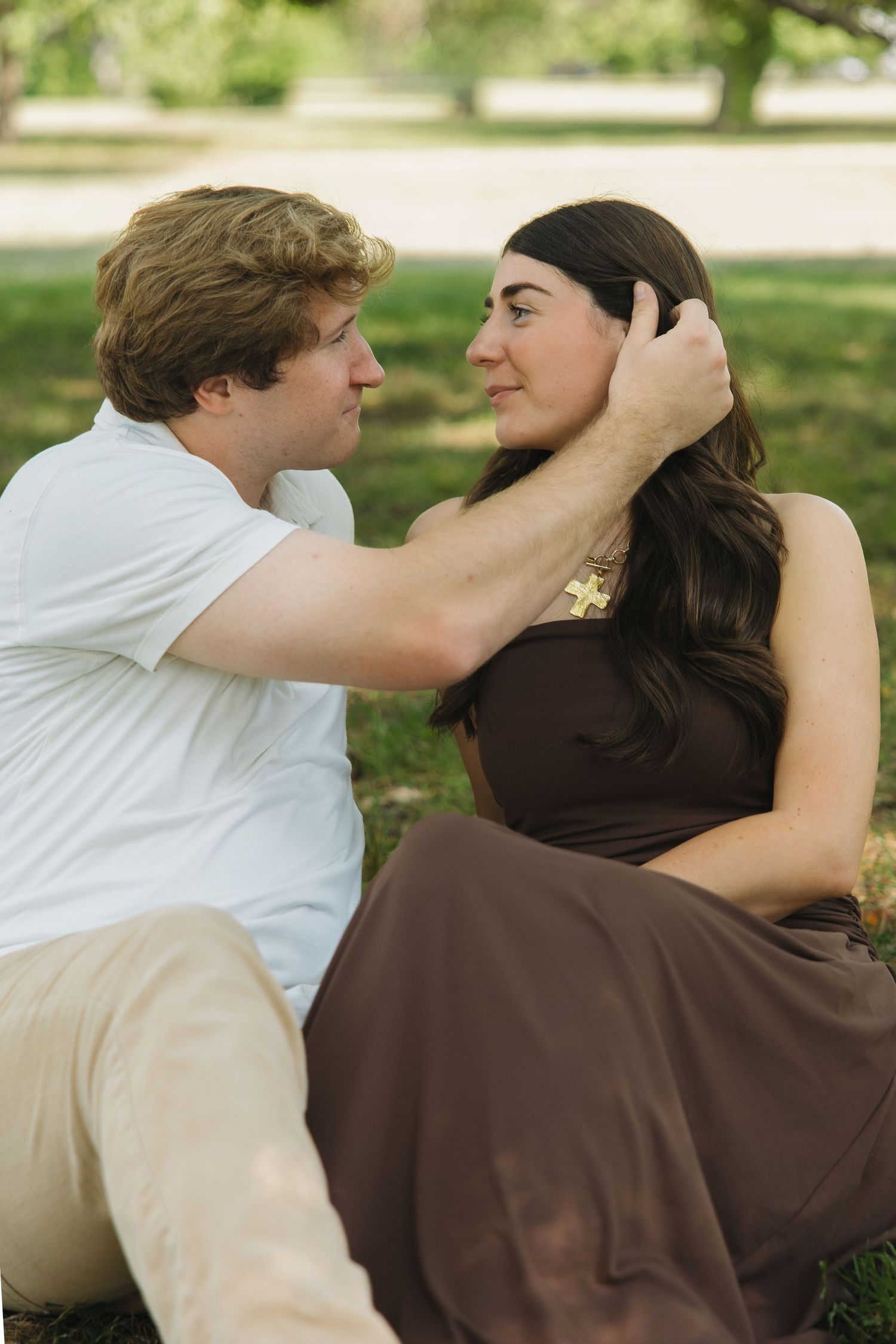 A couple sits on green grass facing each other, man gently touching womans hair in a sunny park.