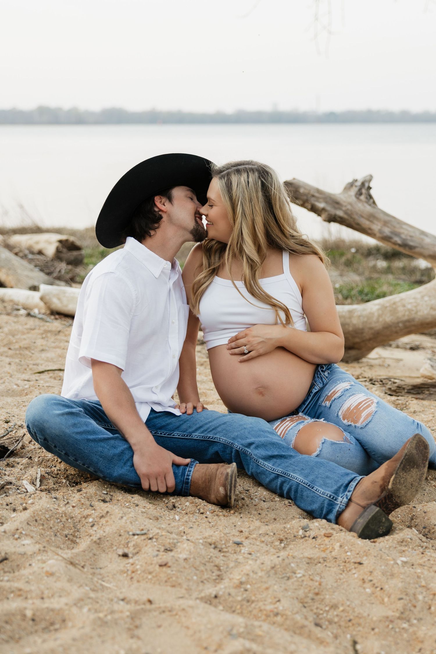 Couple sharing a kiss while sitting on sandy lakeshore, pregnant woman in ripped jeans with partner in cowboy hat.