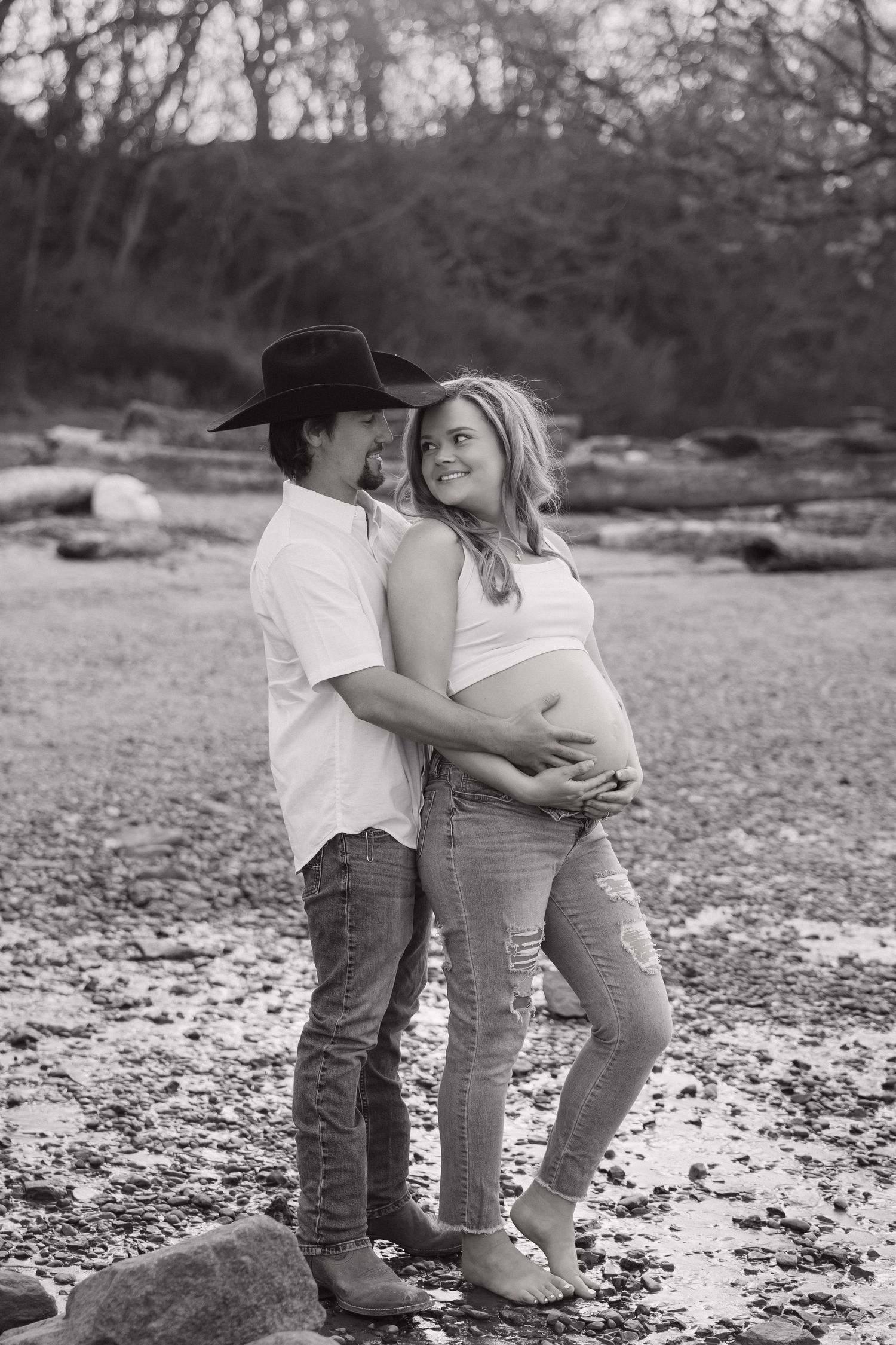 Black and white photo of pregnant couple embracing on rocky beach, man in cowboy hat holding woman's baby bump.