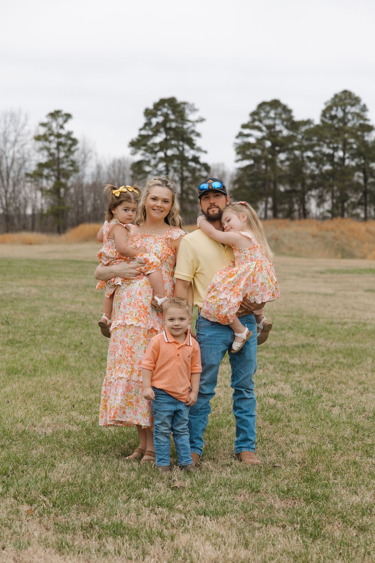 Family of five posing in green field, parents holding two daughters in matching floral dresses, son in orange polo.