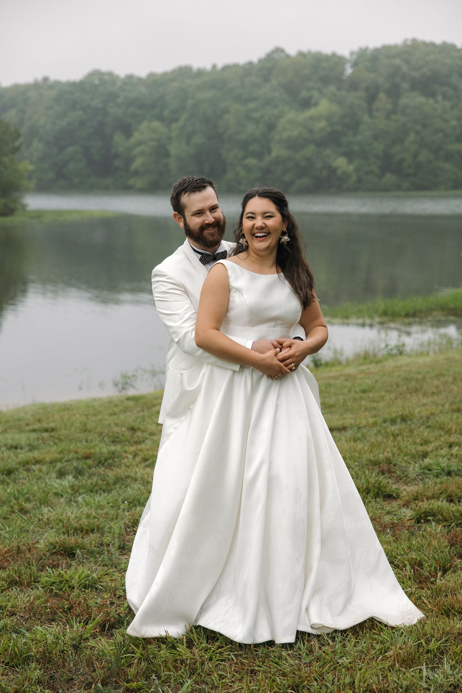 Bride and groom smiling and embracing on a grassy lakeside with misty forested hills behind them.