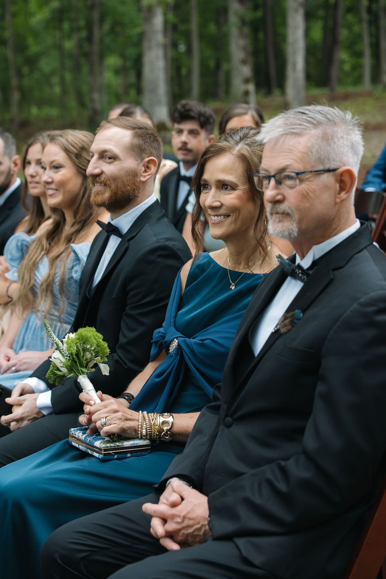 Smiling wedding guests in formal attire seated at outdoor woodland ceremony, woman in teal dress.