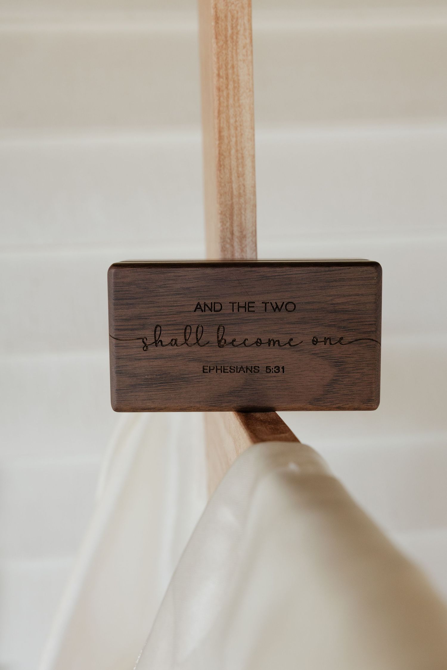 Wooden ring box lid engraved with And The Two Shall Become One Ephesians 5:31 displayed on wooden cross with white draping.