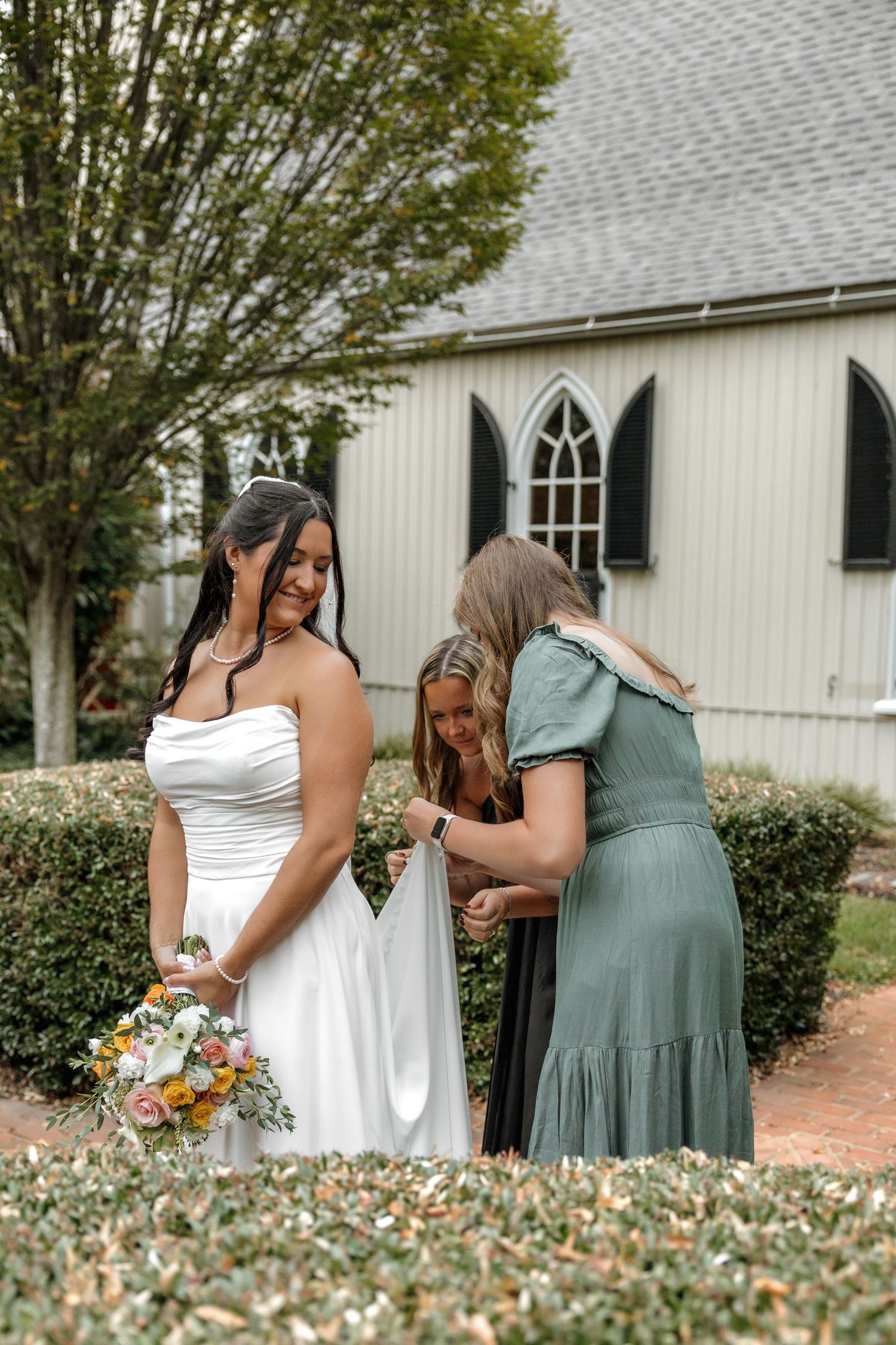 A bride in a white strapless gown holds a bouquet as attendants adjust her dress outside a chapel.