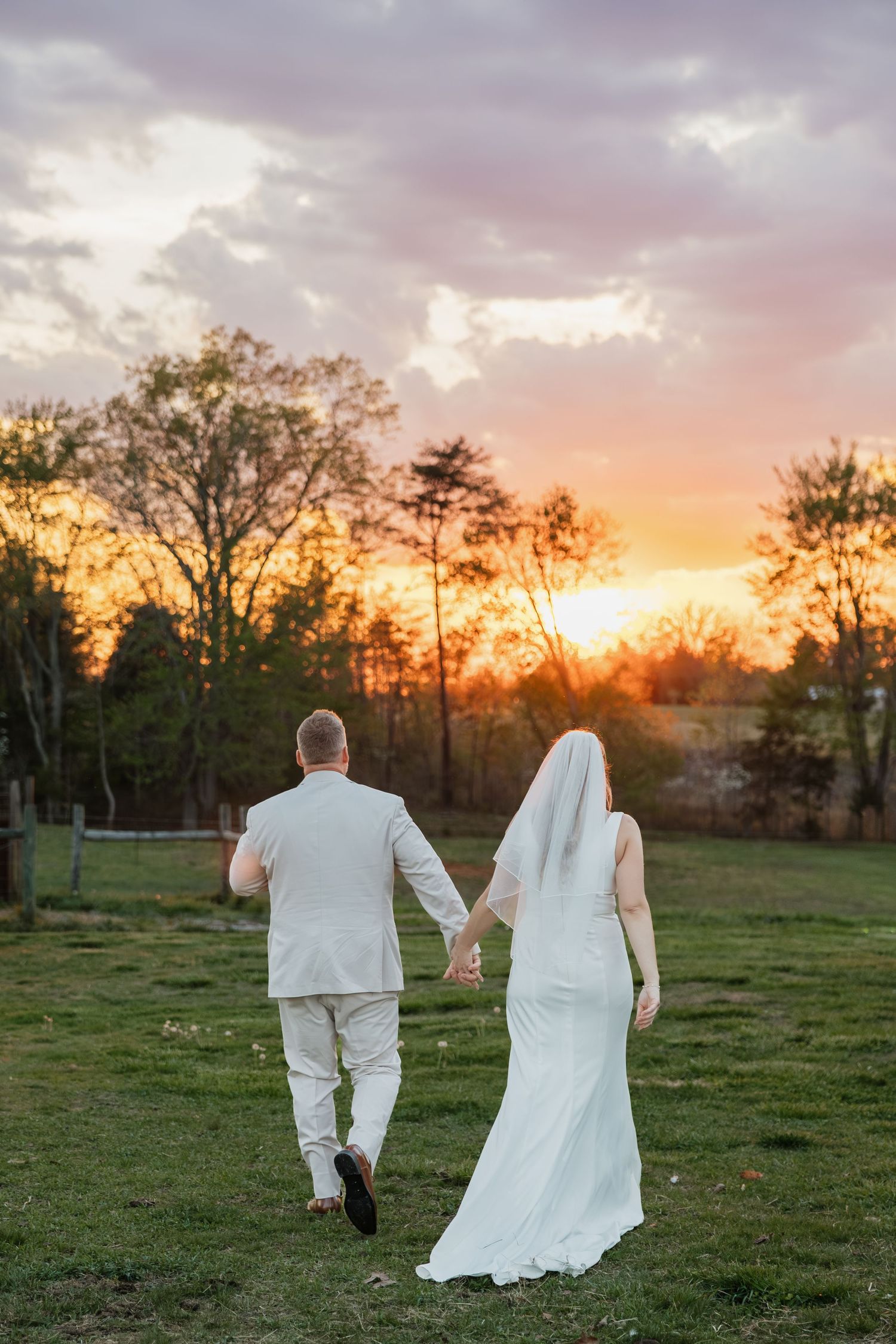 Bride and groom holding hands walking toward a vibrant sunset in a pastoral field.