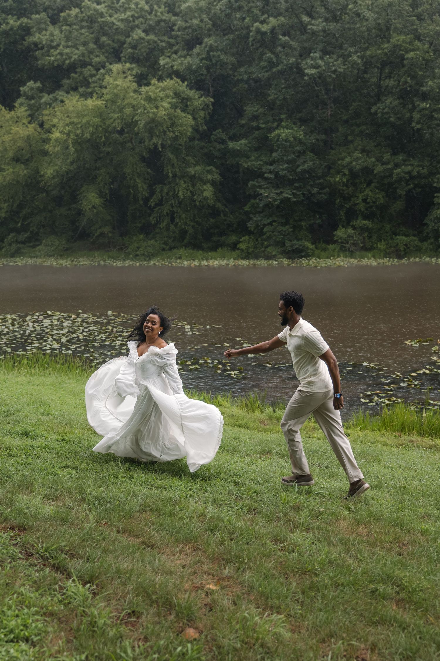Bride in flowing white dress runs playfully by a lake as groom chases her on lush green grass.