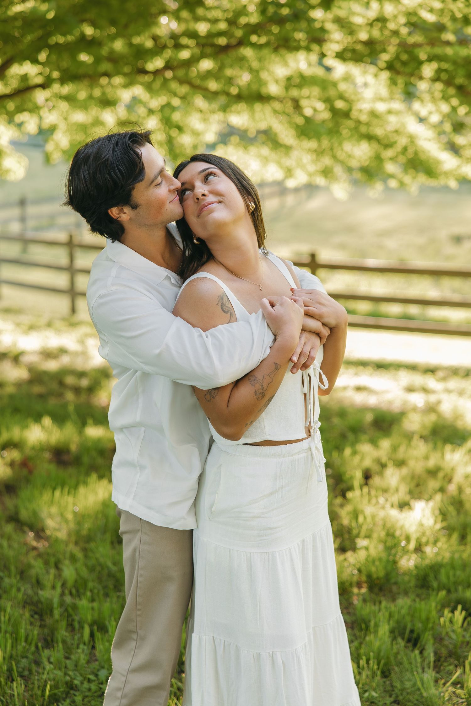 Man tenderly embracing woman in white dress in a sunlit green meadow with wooden fence.
