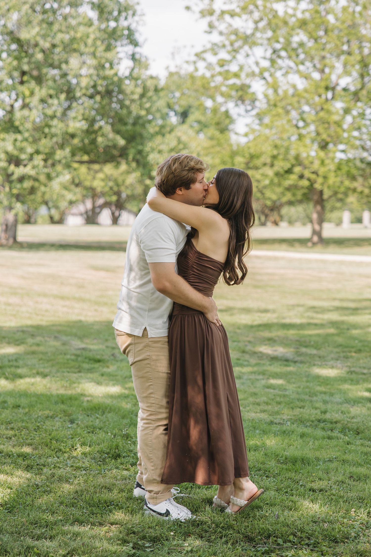 A couple shares a passionate kiss while embracing on a sunny green lawn surrounded by tall trees.