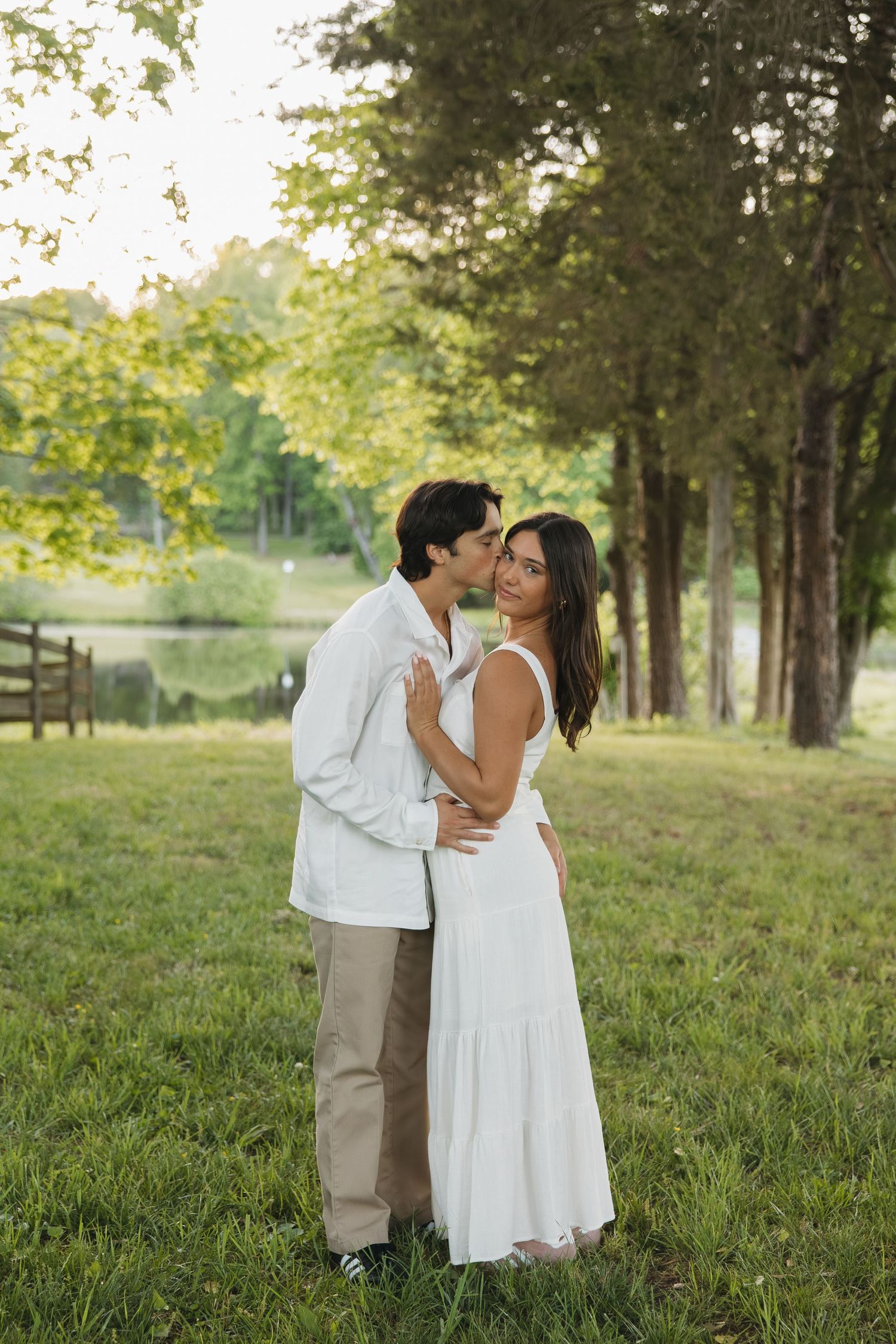A man in white shirt kisses a woman in a white dress on the cheek outdoors near a scenic lake.