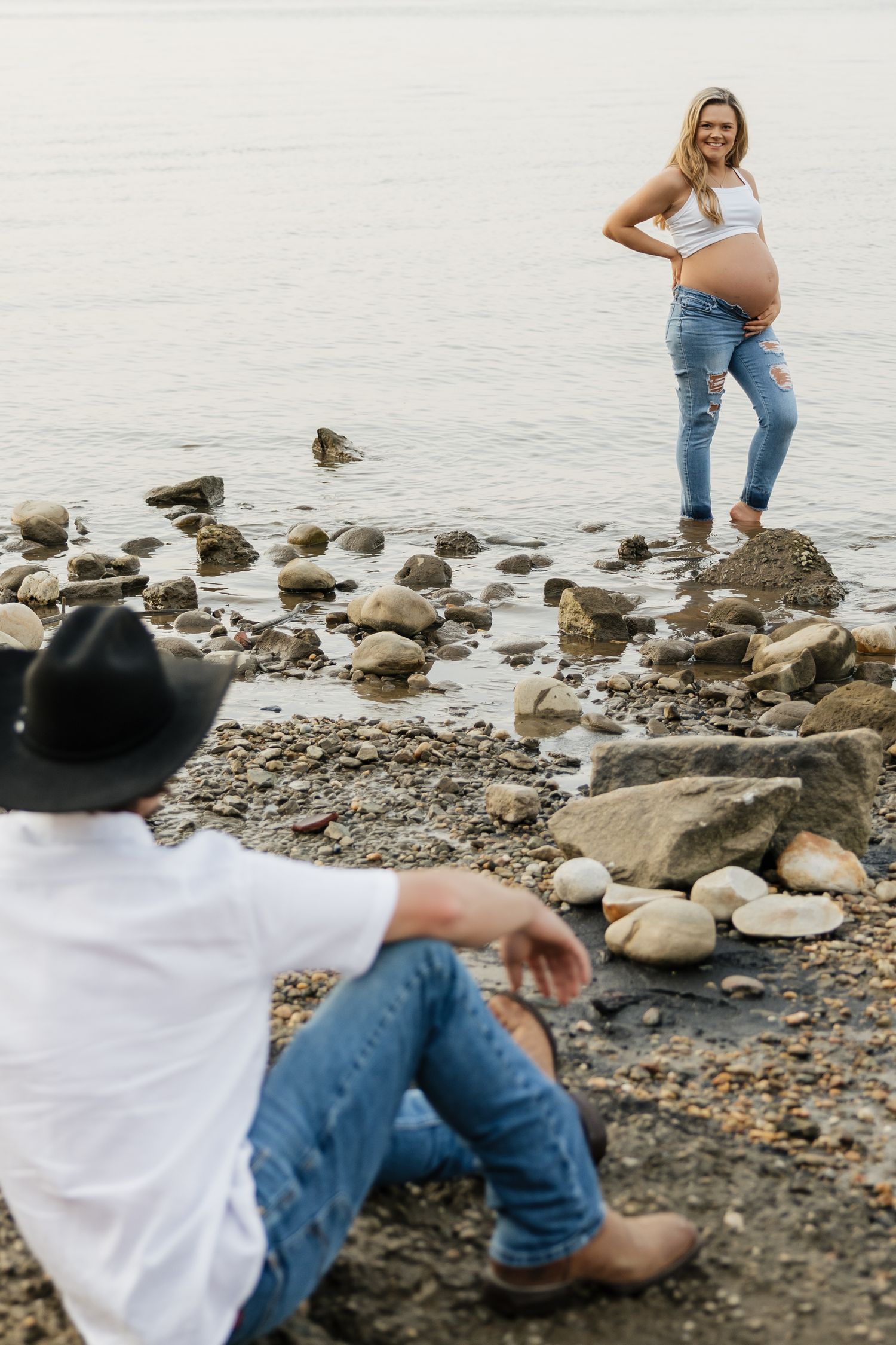 Pregnant woman standing on rocks by river smiling while partner sits on ground watching her.