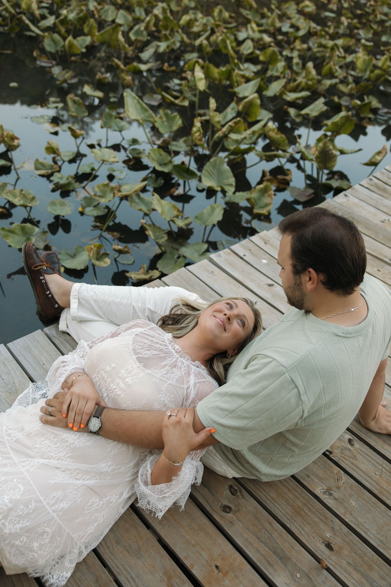 Couple relaxing on a wooden dock, woman in white lace dress lying down while man sits beside her near lily pads.