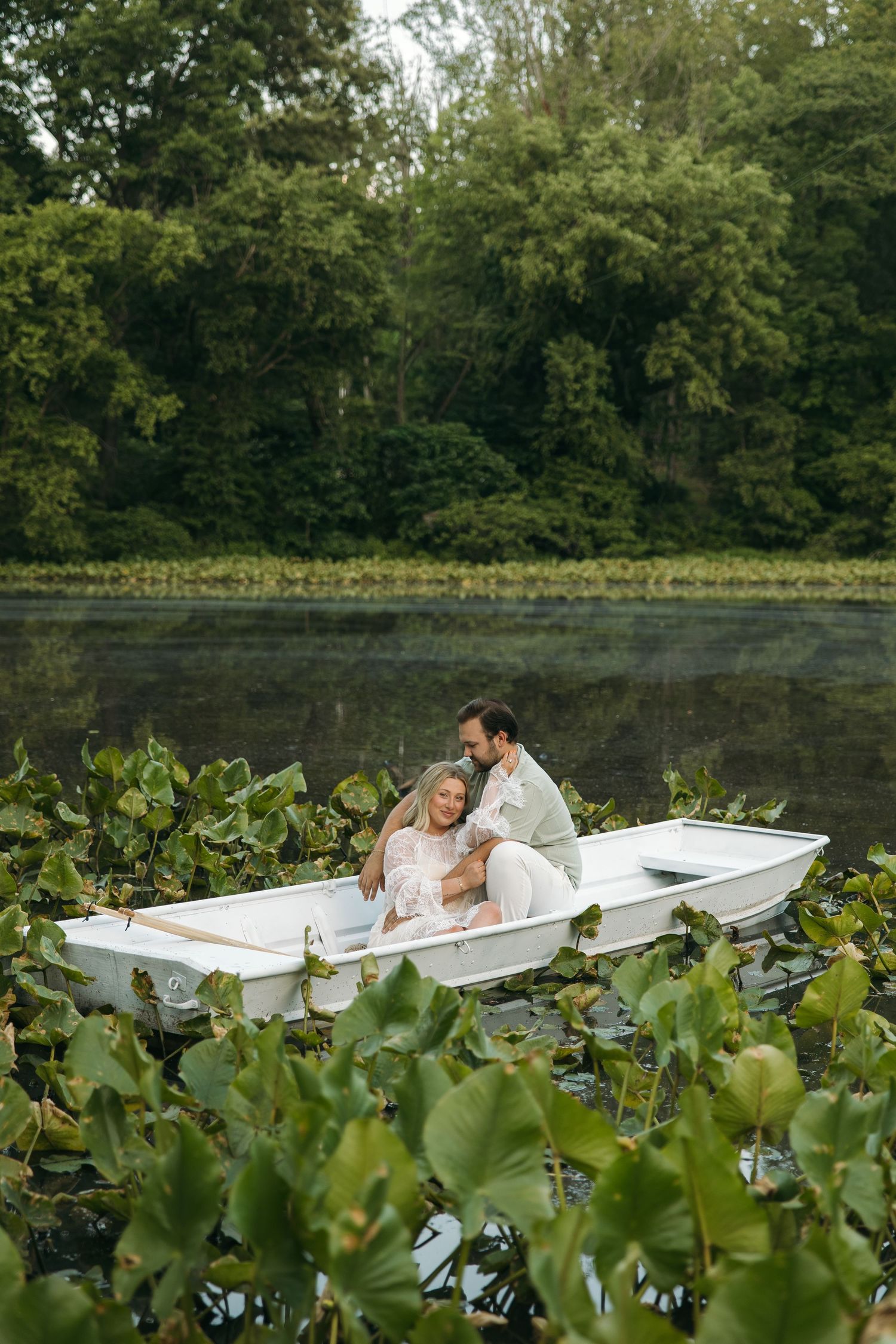 Romantic couple embracing in a white boat amid lily pads on a calm lake surrounded by dense greenery.