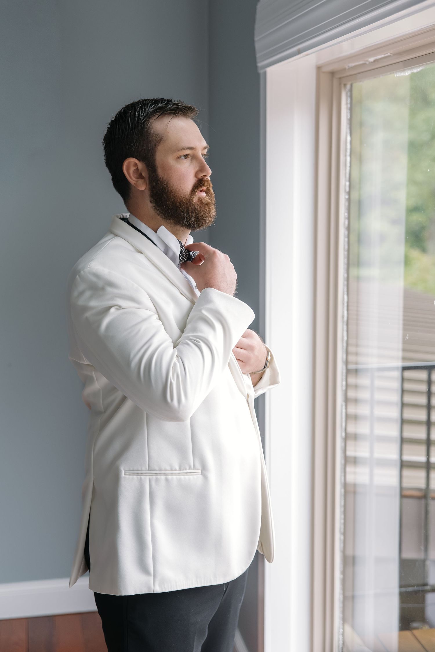 Handsome bearded groom in white blazer adjusting bow tie while gazing out window before wedding.