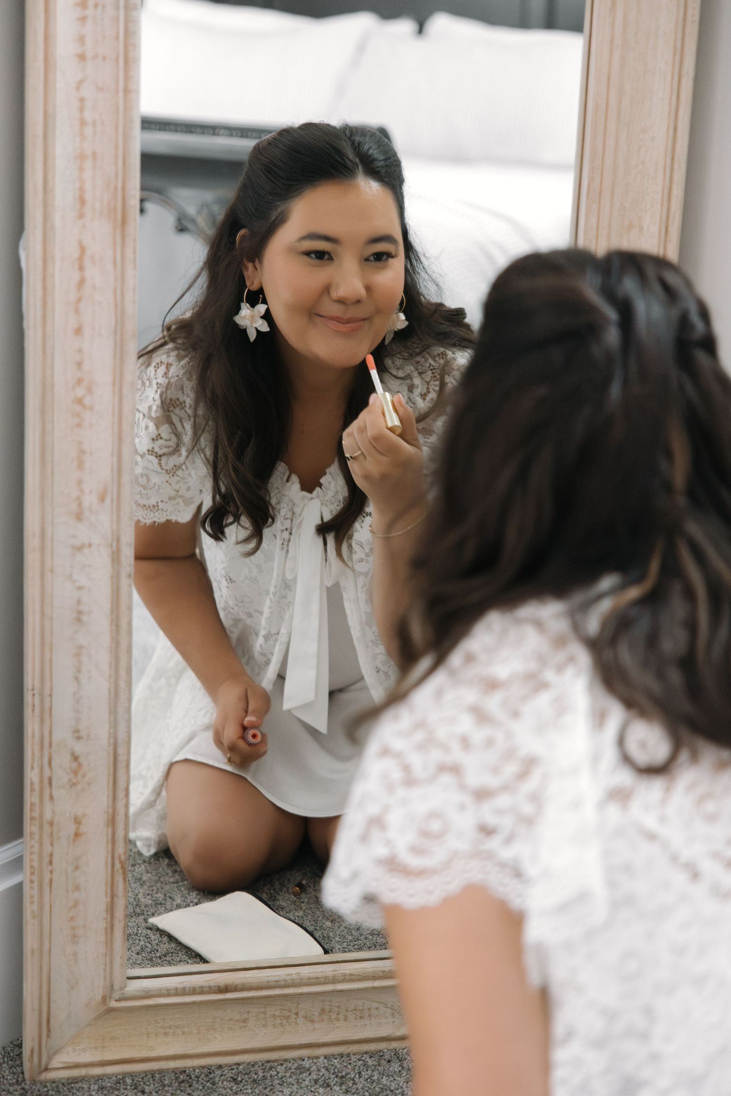 Smiling bride in white lace dress applying lipstick while looking in an ornate floor mirror.