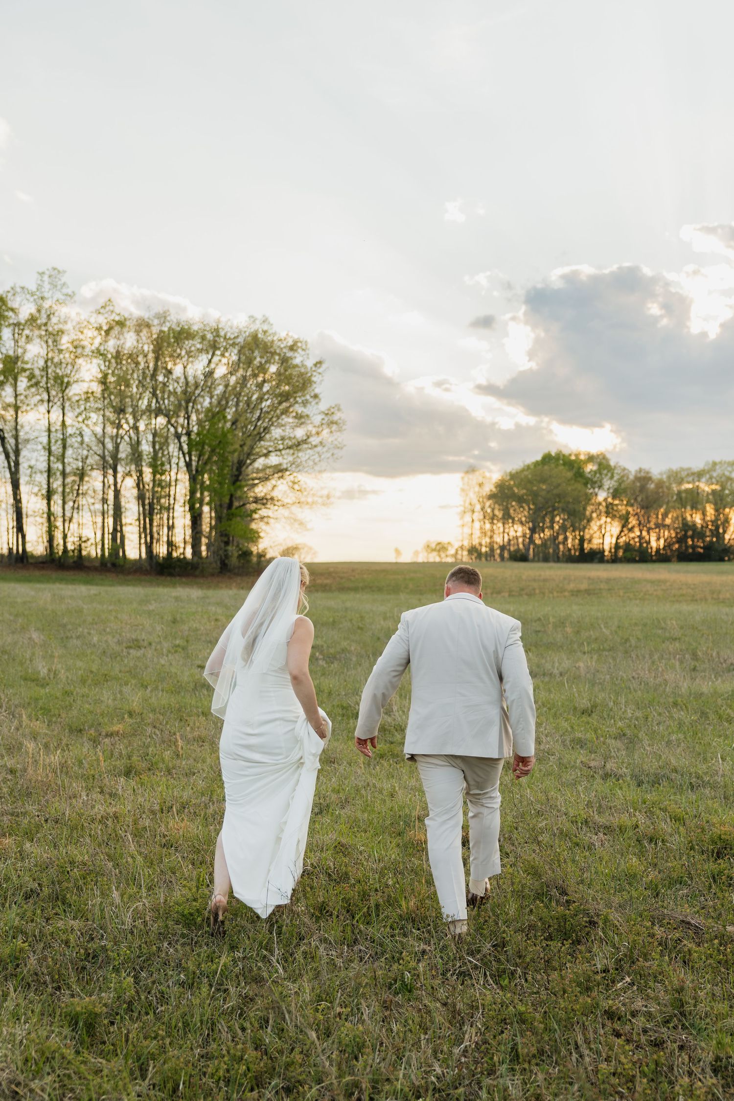 Bride and groom walk hand in hand through a green field toward a stunning sunset.