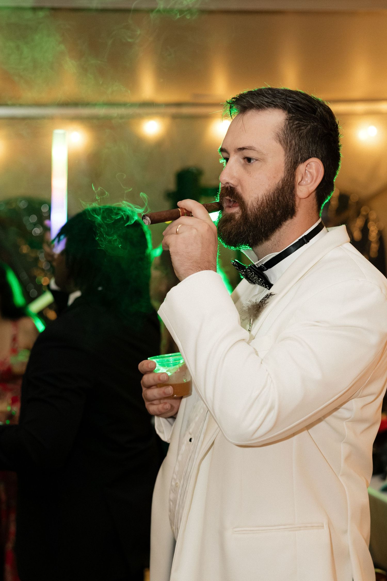 Dapper groom in white tuxedo jacket smoking a cigar and holding a drink at wedding reception.
