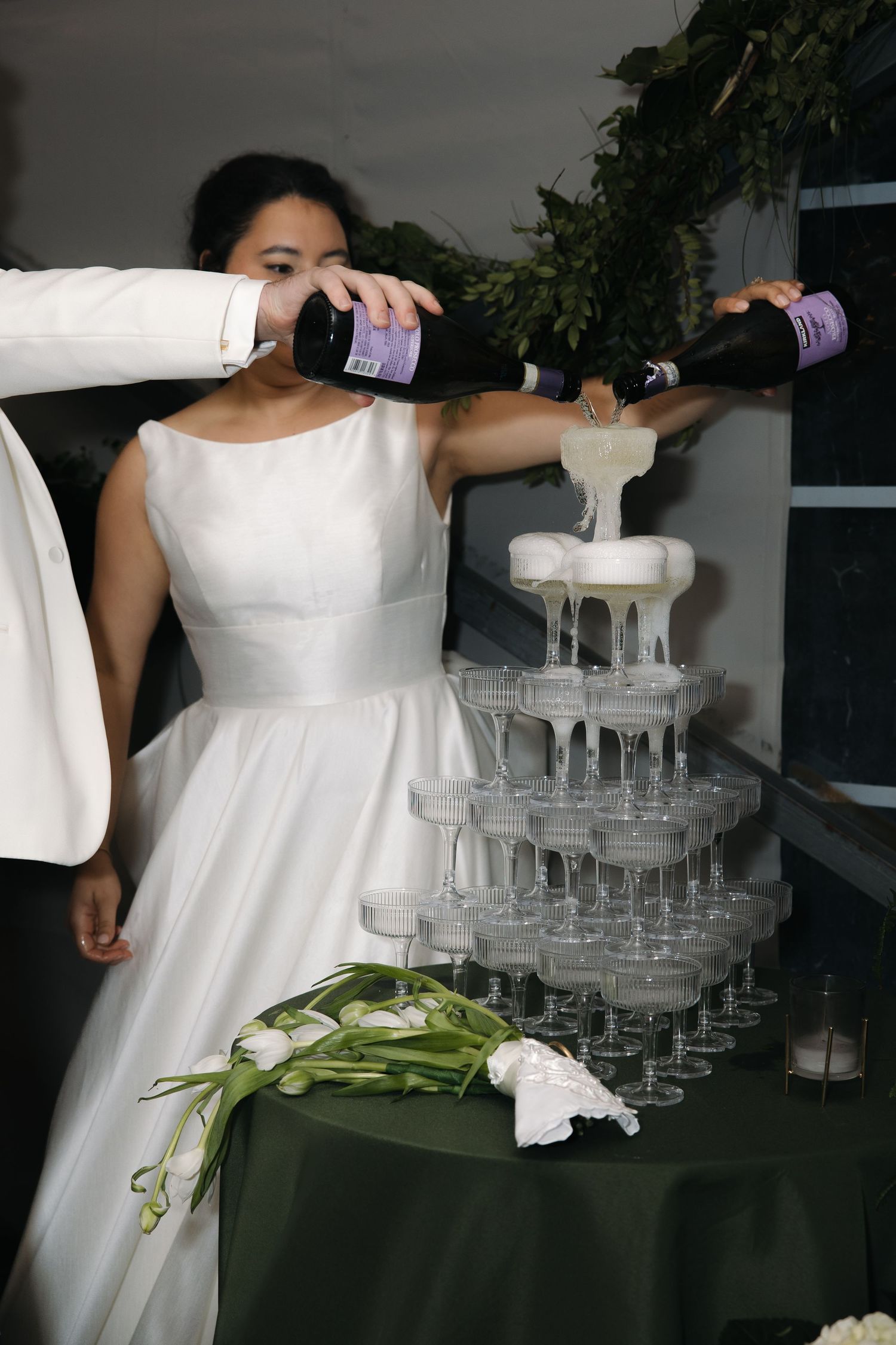 Bride in white gown and groom pouring champagne into elegant tiered glass tower at wedding.