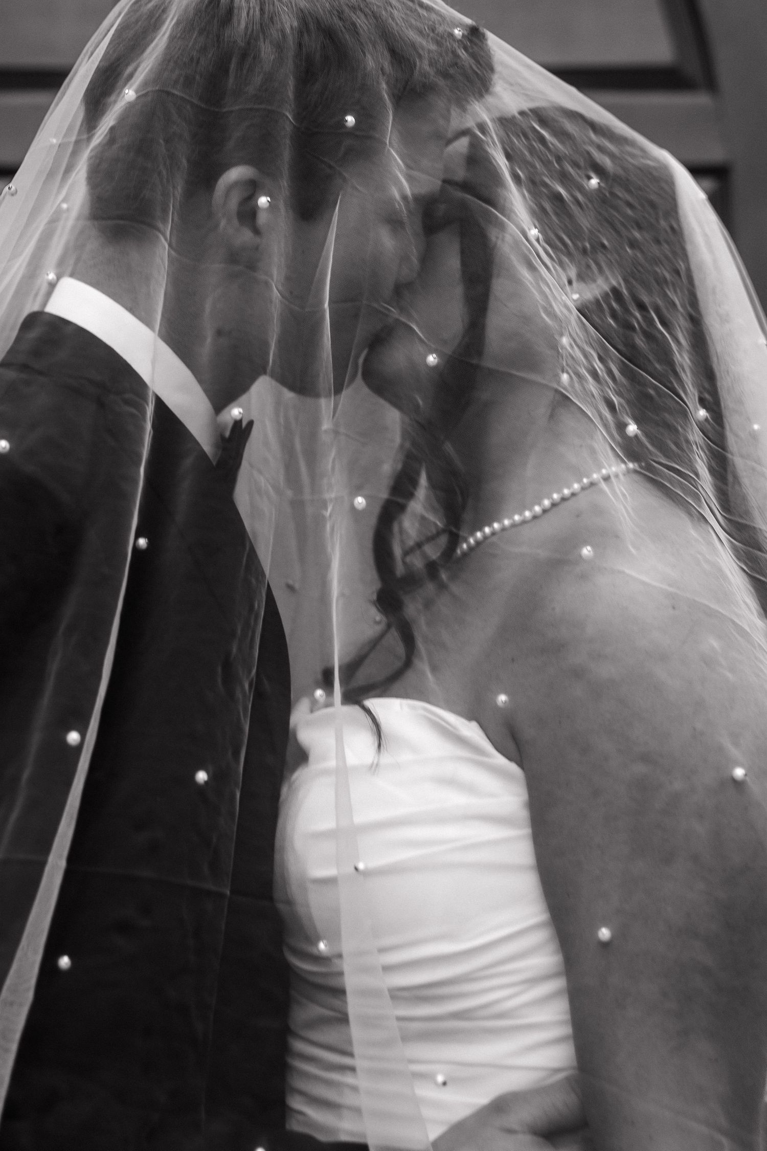Black and white romantic photo of bride and groom kissing beneath a pearl-adorned wedding veil.