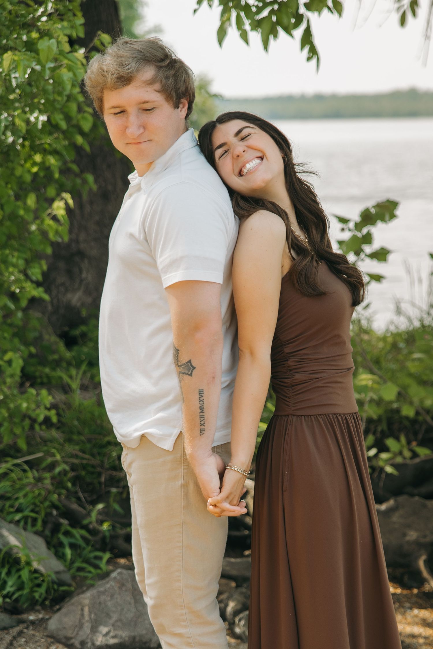 Couple standing back-to-back near a lake, woman in brown dress laughing joyfully while holding hands.
