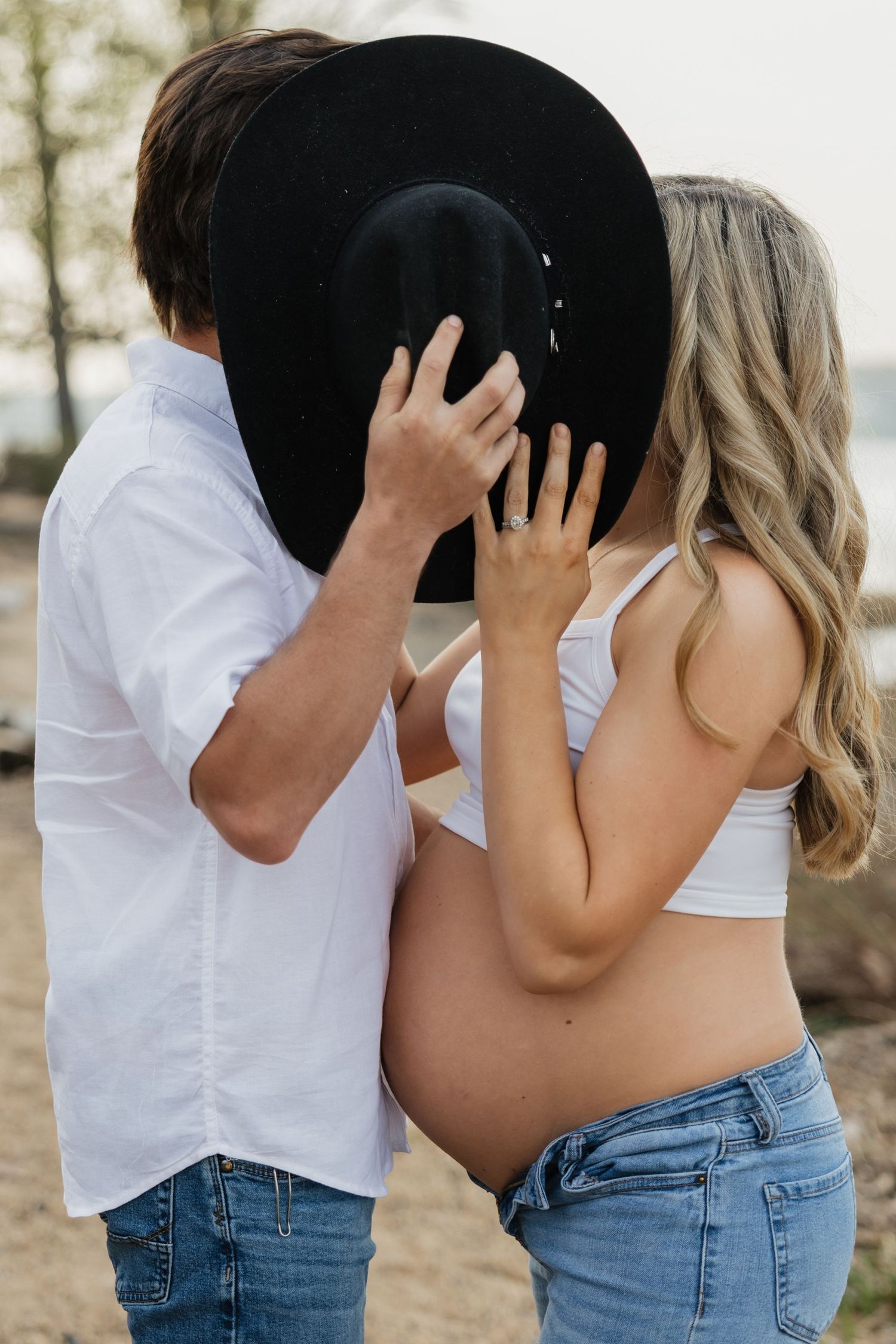 Pregnant couple shares a kiss hidden behind a black cowboy hat on a beach, woman showing baby bump.