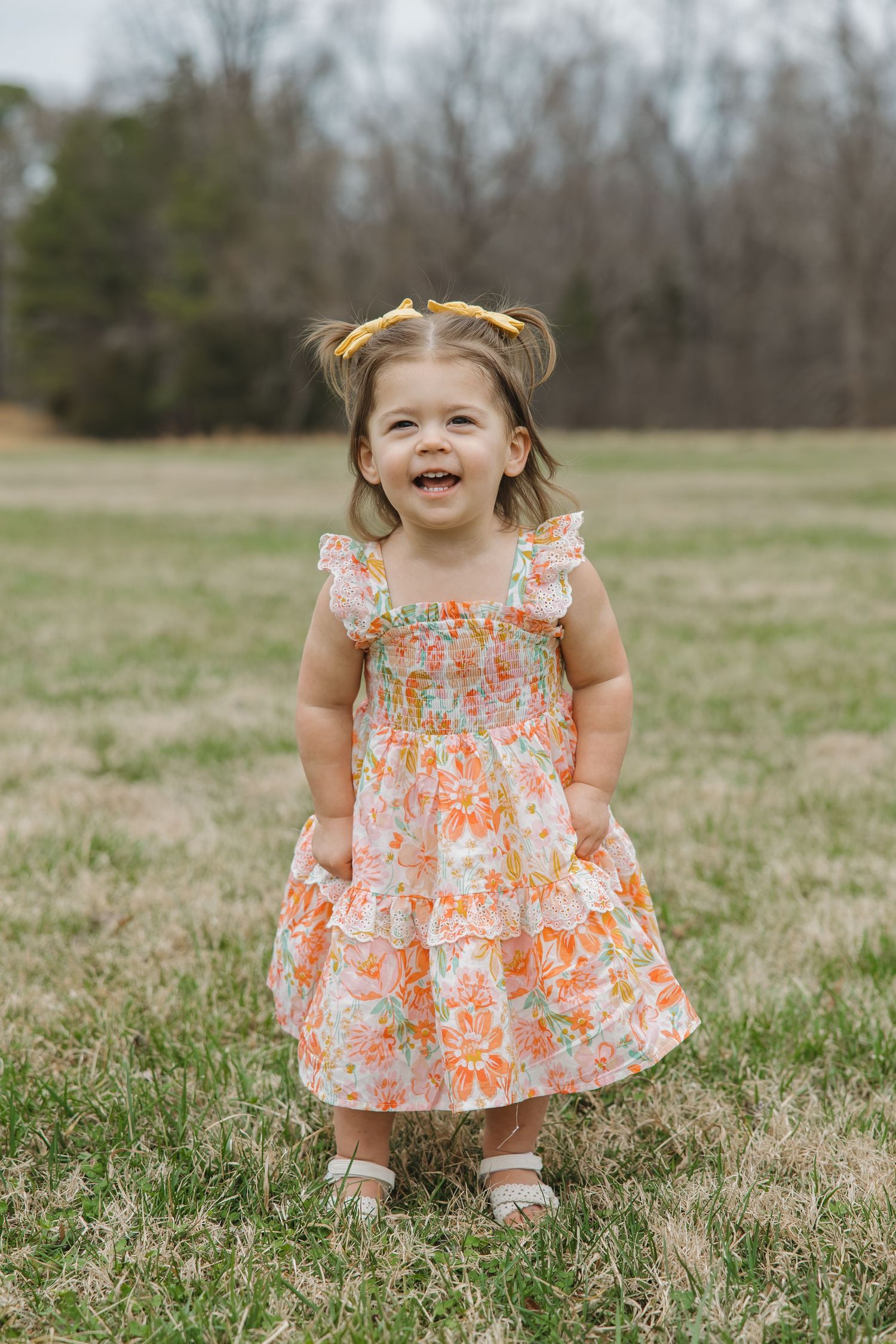 Adorable toddler girl in orange floral dress with yellow hair bows laughs joyfully while standing in a grassy field.