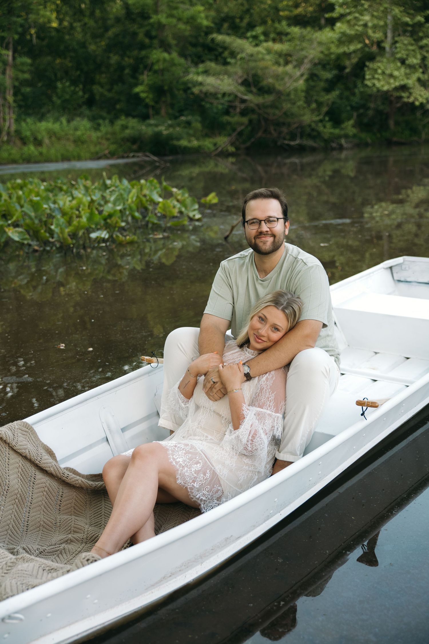 Couple sharing a tender embrace in a white rowboat surrounded by lush greenery on a calm river.