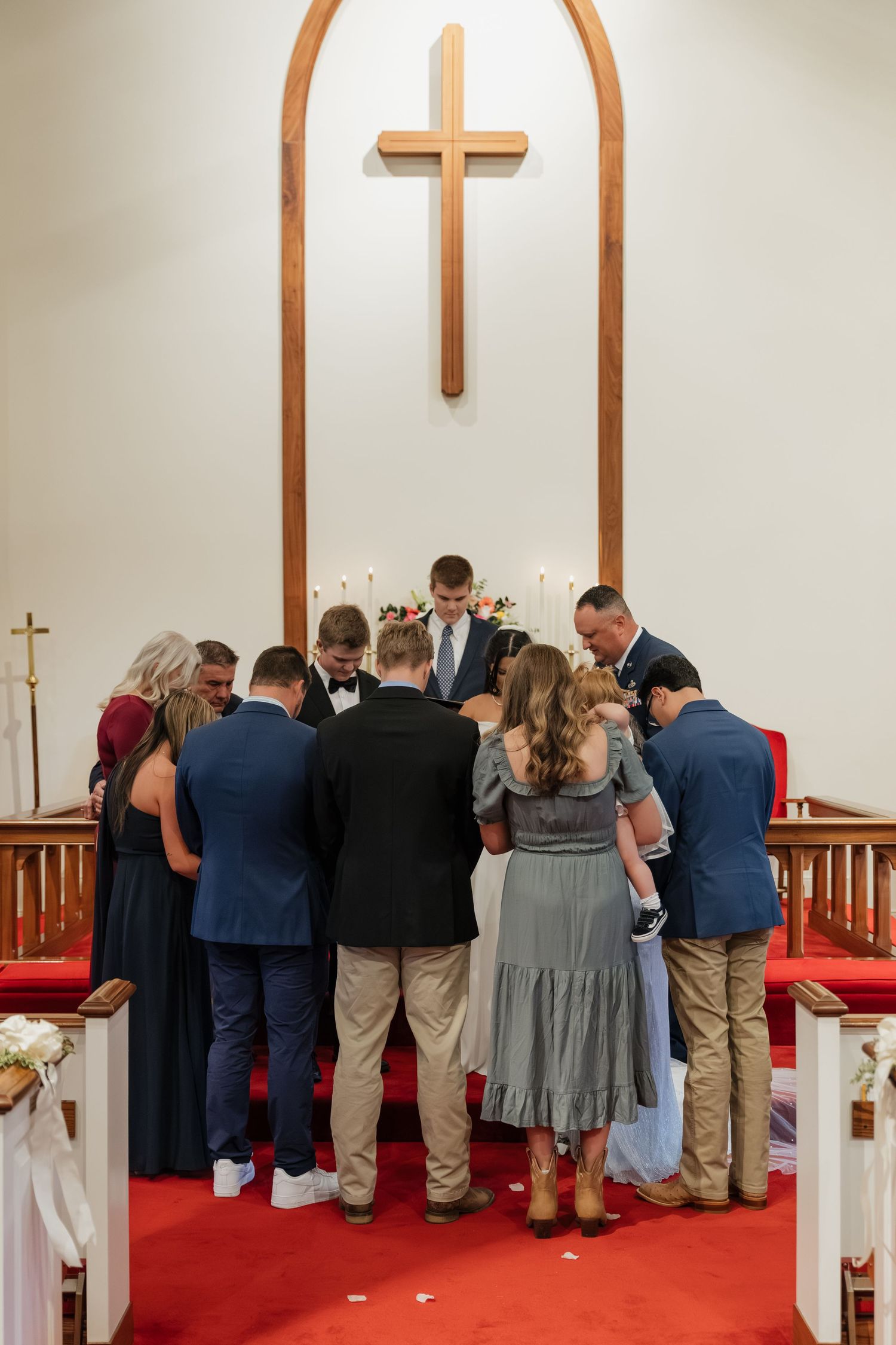 Wedding party gathered in a circle for prayer beneath a wooden cross at the church altar.