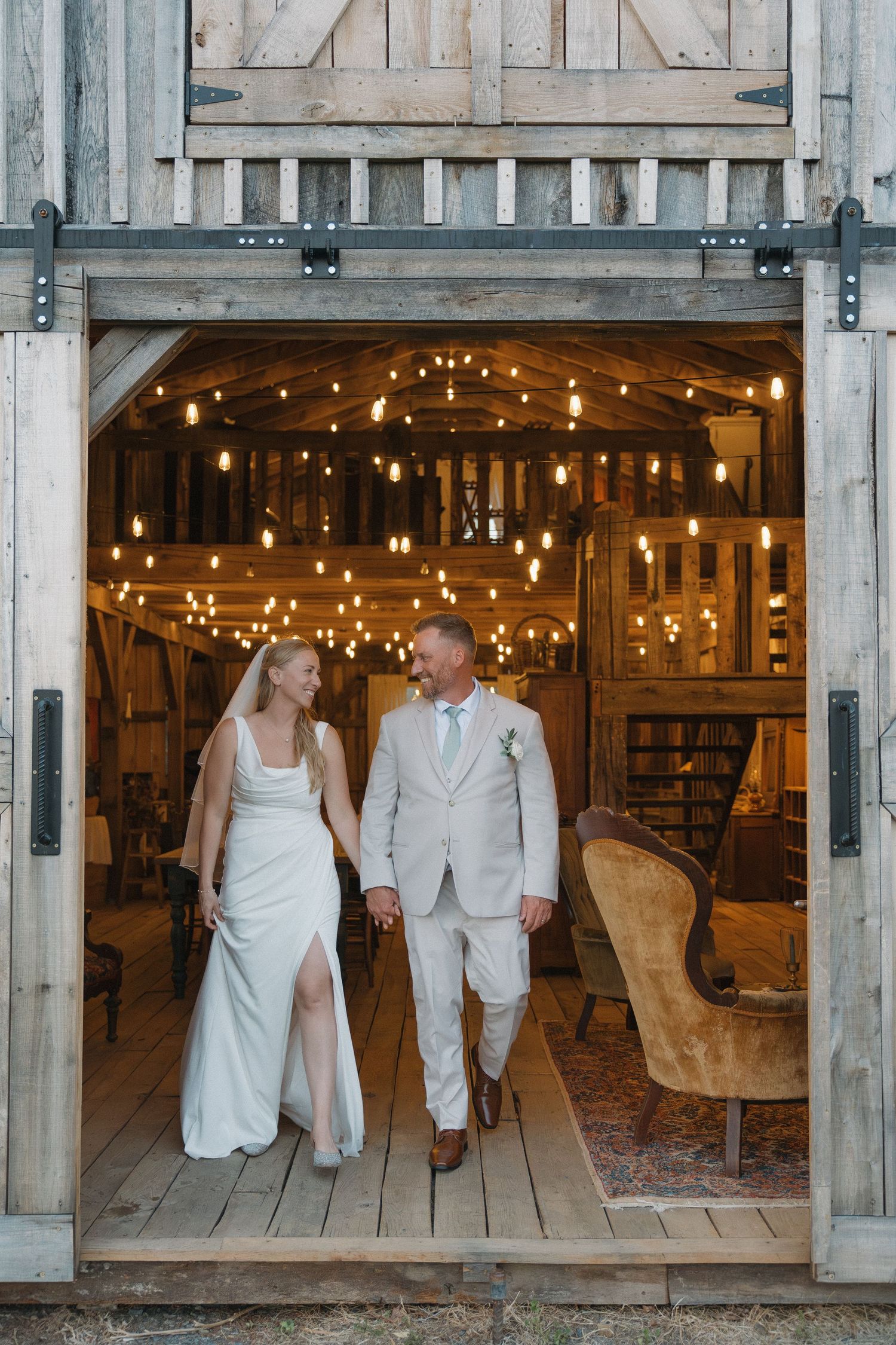 Bride and groom walk hand-in-hand through rustic barn doors adorned with warm Edison bulb string lights.