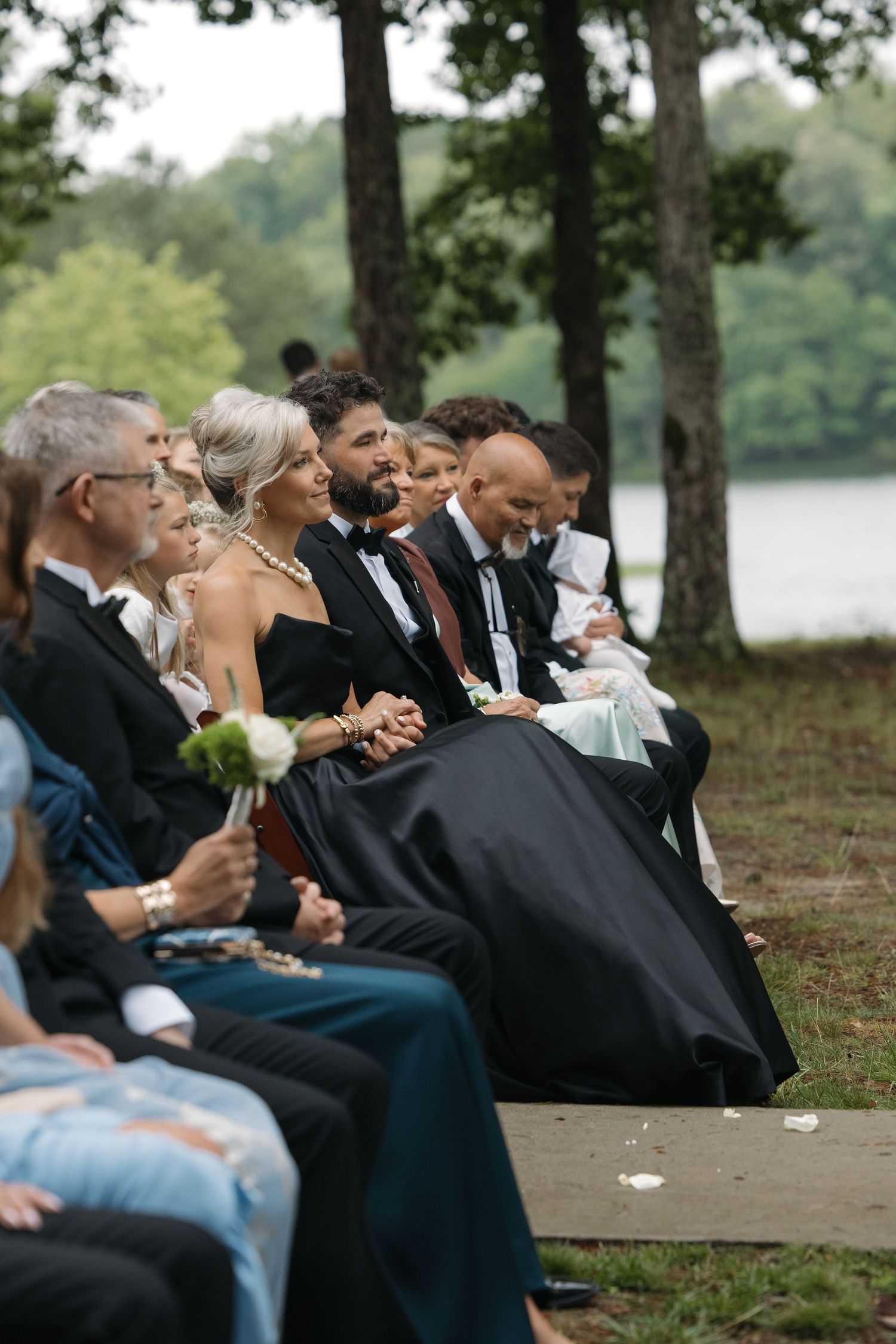 Wedding guests seated outdoors near a lake, dressed in formal attire, watching the ceremony with smiles.
