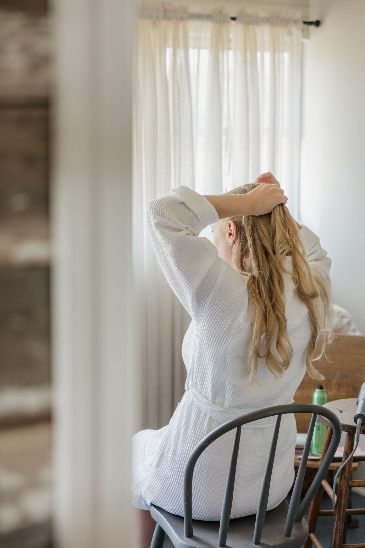 Blonde bride in white robe styling her hair while seated at window-lit vanity table.