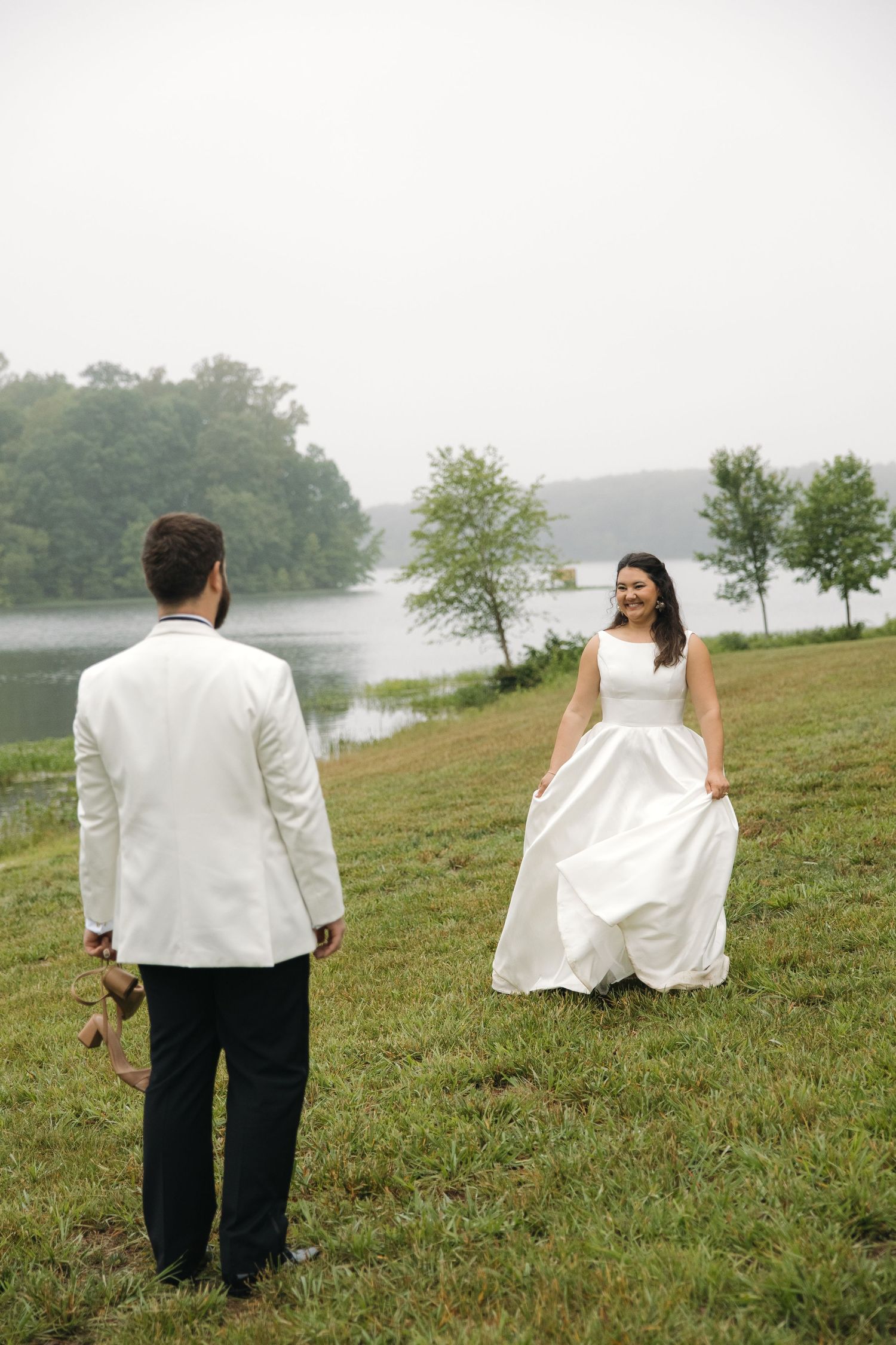 Bride and groom face each other on grassy lawn with foggy lake behind.