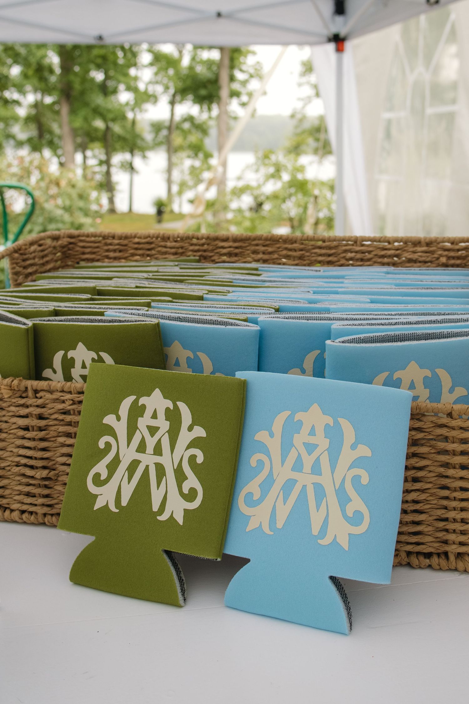 Monogrammed wedding koozies in olive green and sky blue displayed in a wicker basket at an outdoor reception.