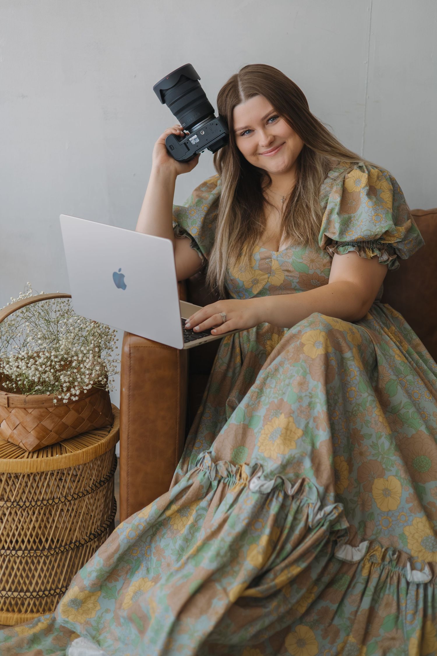 Woman in floral dress holding a camera and using a MacBook laptop while seated on a leather chair.