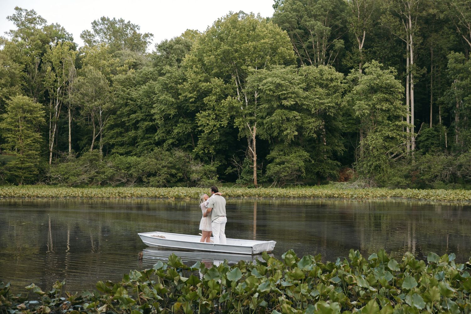 Couple standing close together in a white rowboat on a serene river surrounded by green trees.