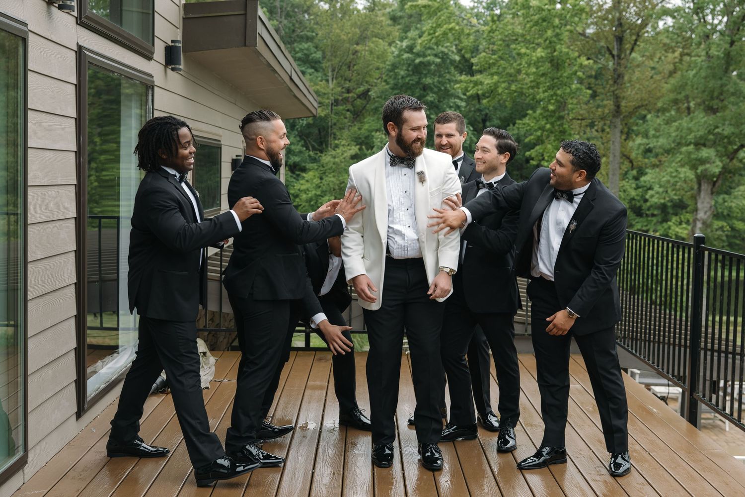 Groom in white tuxedo jacket celebrates with groomsmen in black tuxedos on a wooden deck outdoors.