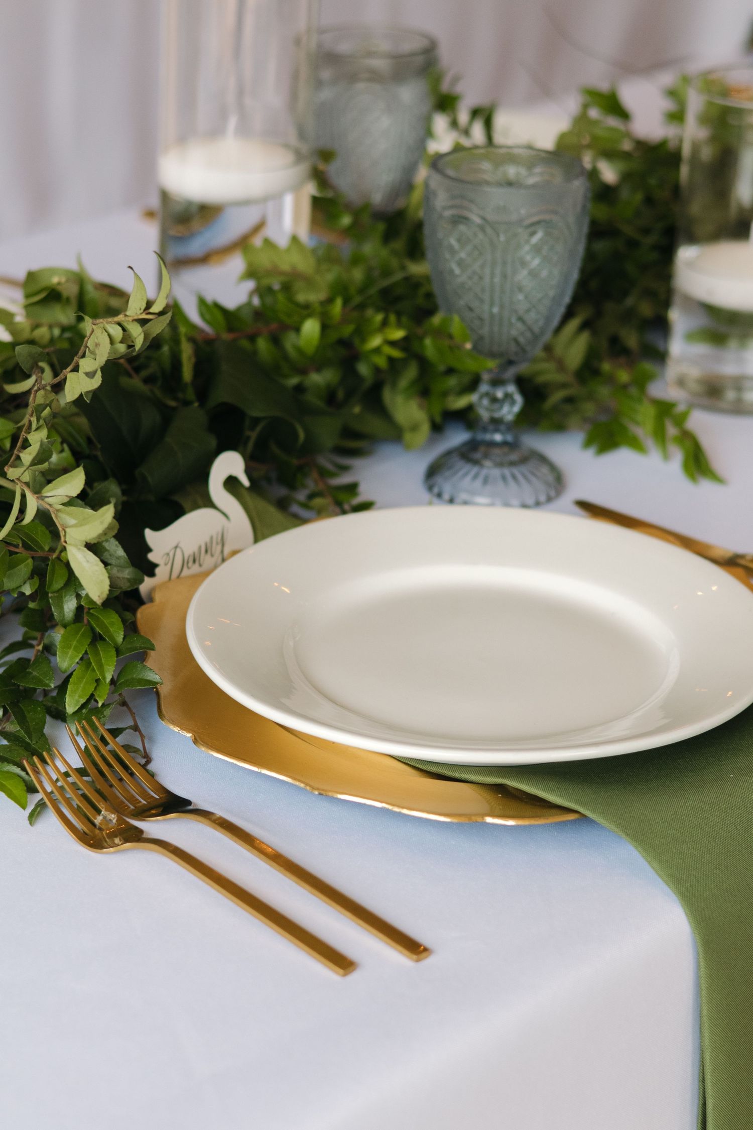 Wedding table setting with gold-rimmed plates, gold cutlery, green napkin, and lush greenery runner.