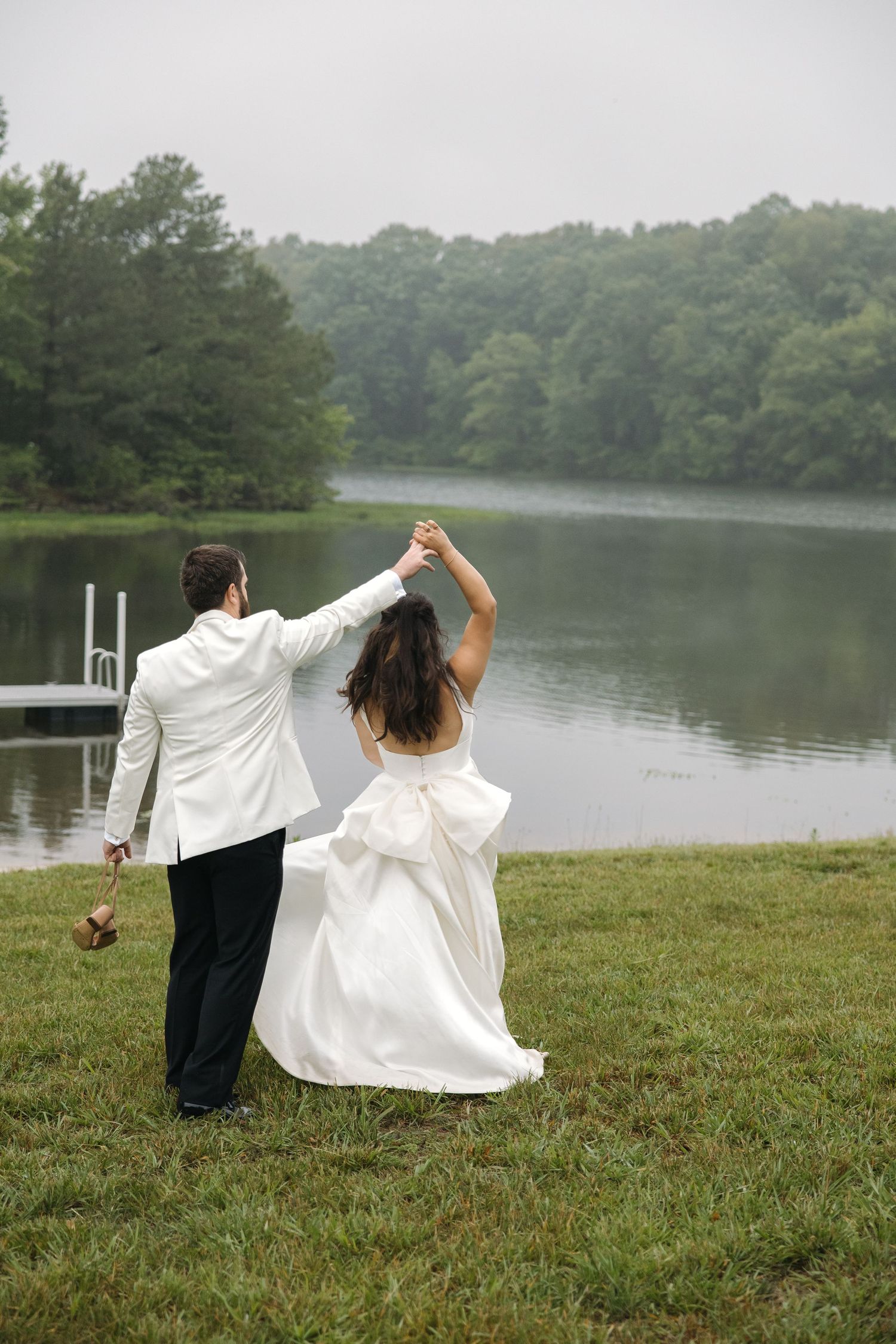 Bride and groom hold hands raised while walking toward misty lake surrounded by green forest.