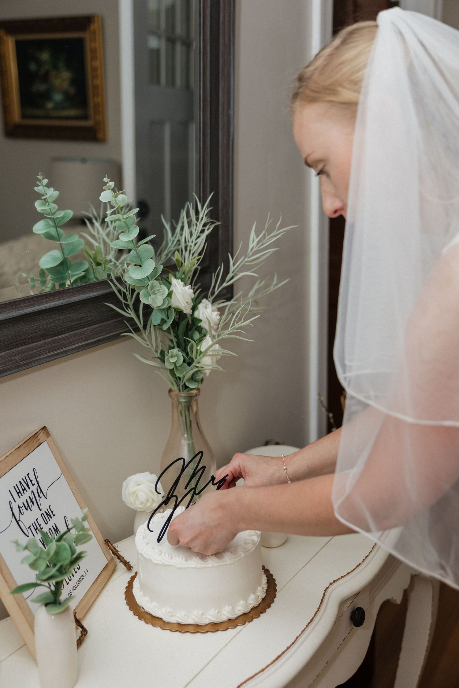 Bride in white veil signs wedding cake decorated with greenery and Mr. and Mrs. calligraphy topper.