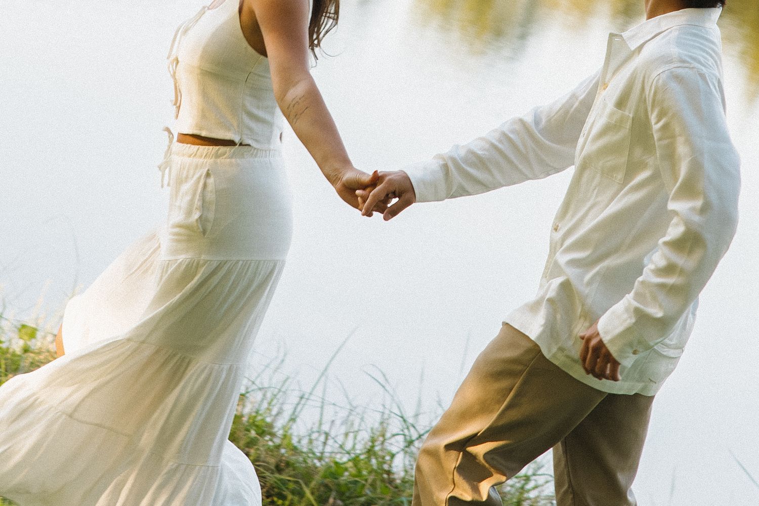 Couple in white outfits holding hands walking along a peaceful lakeside with tall grass and calm water.