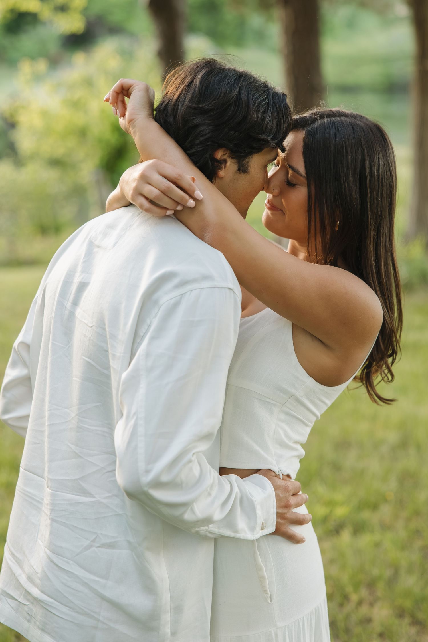 Couple in white outfits sharing a tender moment, nearly kissing in a green outdoor setting.