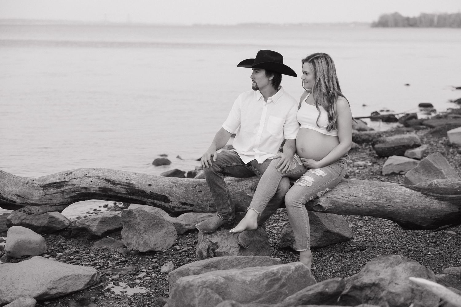 Black and white maternity photo of couple sitting on rocks by the water, woman displaying pregnant belly.