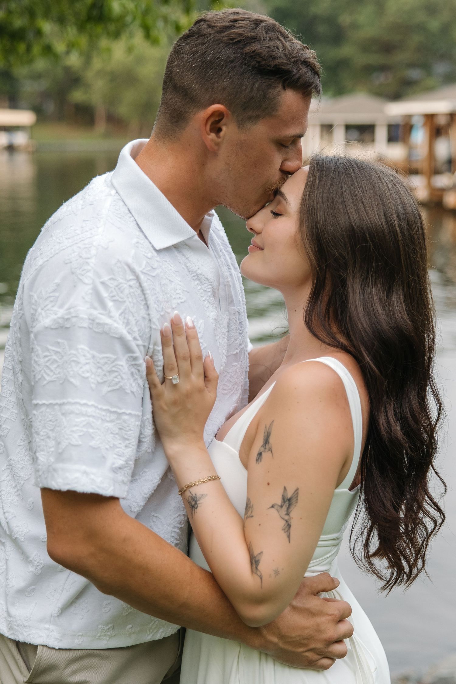 Couple in white outfits sharing a romantic kiss near a lake, woman with tattoos wearing an engagement ring.