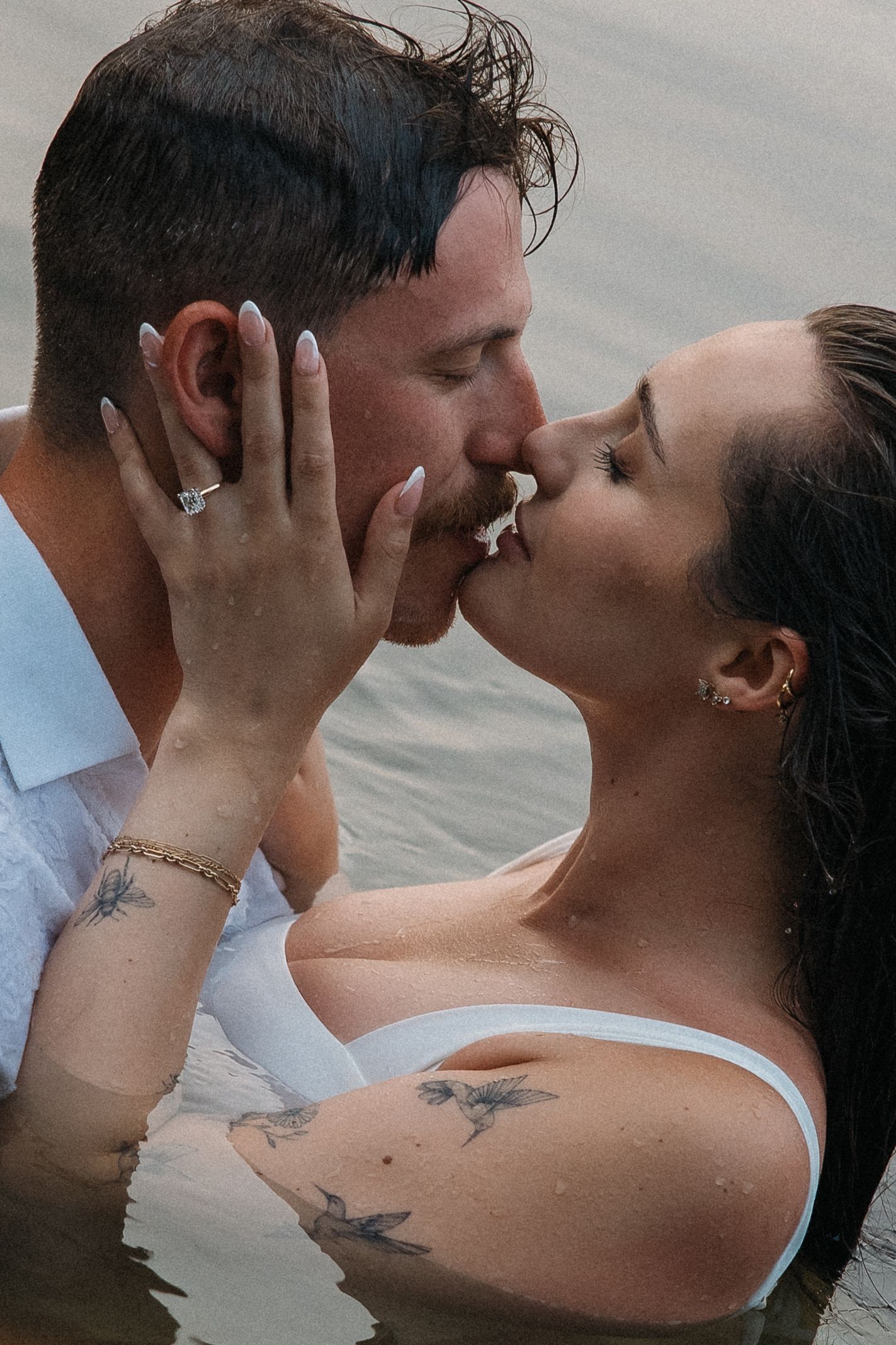 Couple sharing a romantic kiss in water, woman with tattoos and engagement ring holding man's face tenderly.