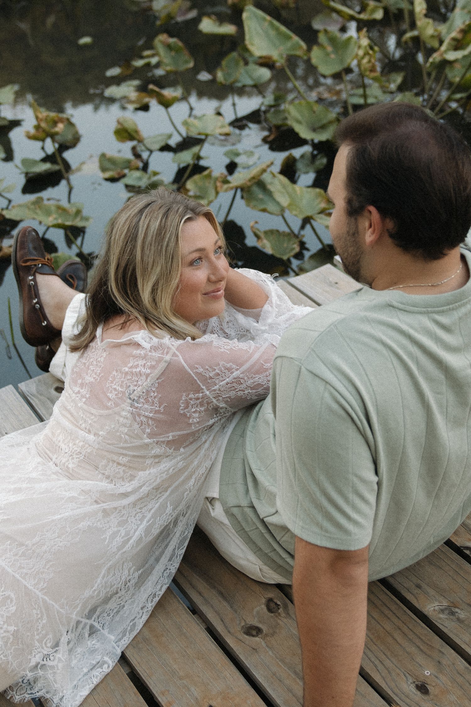 Woman in white lace dress lying on a wooden dock smiling at her partner near a lily pad pond.