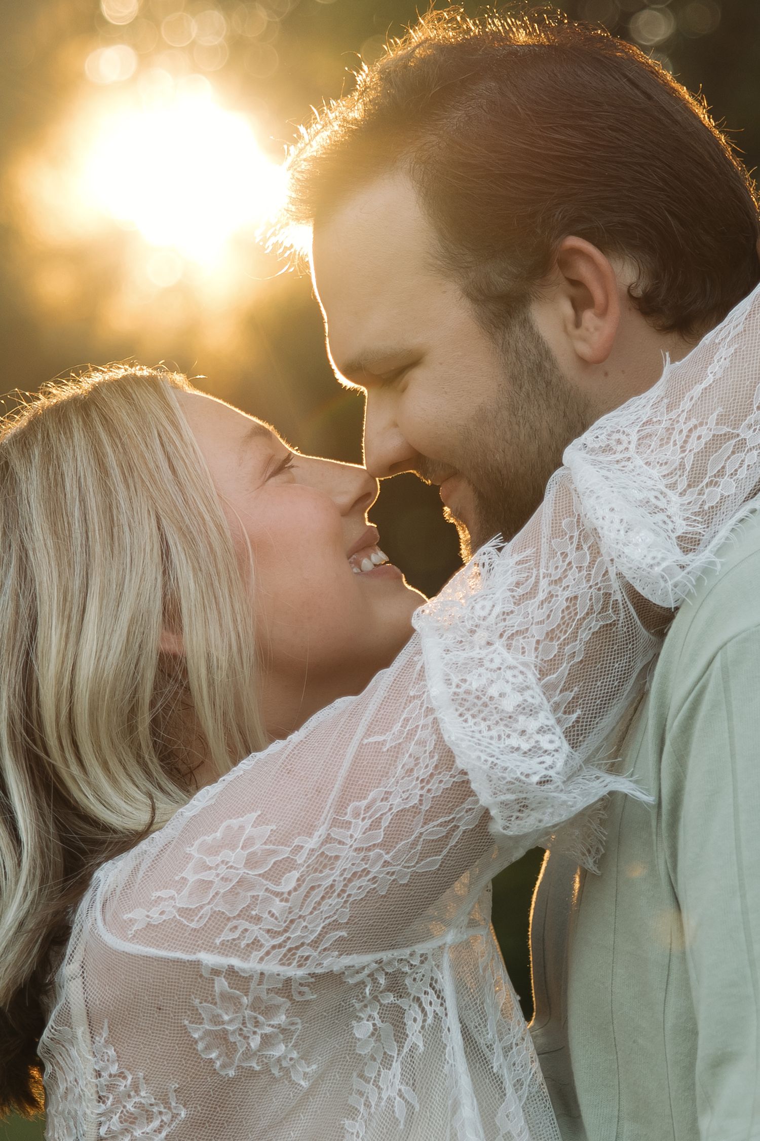 Romantic couple nearly kissing at golden sunset, woman in white lace dress embracing man with warm sun flare behind them.
