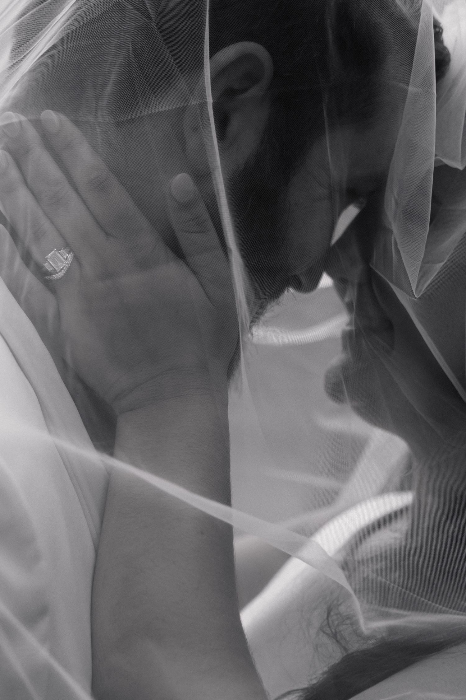 Intimate black and white wedding photo of couple kissing under bridal veil with ring visible.